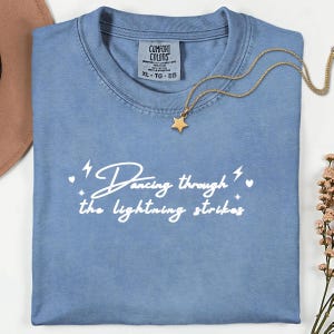 Pode incluir: Uma camiseta azul clara Comfort Colors com a frase "Dancing through the lightning strikes" em branco. Um colar com pingente de estrela dourada repousa sobre a camiseta. A camiseta está dobrada.