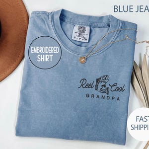 May include: Blue jean colored embroidered shirt with the text "Reel Cool Grandpa" and a fish graphic. The shirt has a round neck and a gold necklace. The words "Embroidered Shirt" are in a circle on the shirt.