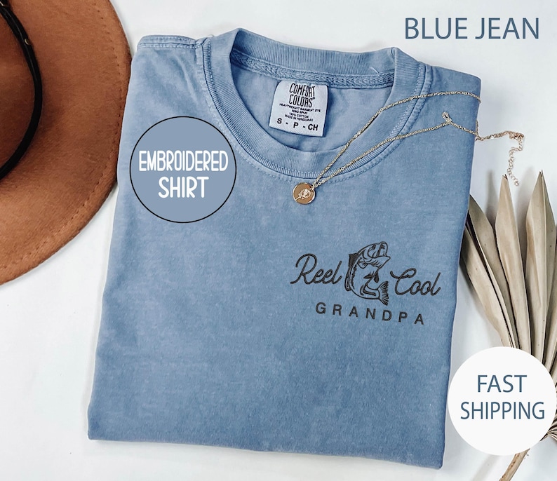 May include: Blue jean colored embroidered shirt with the text "Reel Cool Grandpa" and a fish graphic. The shirt has a round neck and a gold necklace. The words "Embroidered Shirt" are in a circle on the shirt.