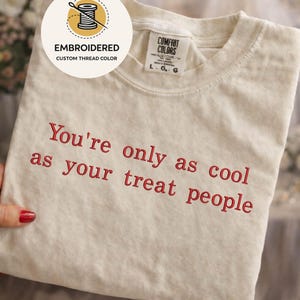 May include: A cream-colored short-sleeve t-shirt with the embroidered red text "You're only as cool as your treat people" on the front. The shirt features a Comfort Colors brand label at the neck and a circular sticker indicating custom thread color.