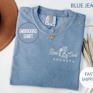 May include: Blue jean colored embroidered shirt with the text "Reel Cool Grandpa" and a fish graphic. The shirt has a round neckline and short sleeves. A gold necklace with a pendant is draped on the shirt.