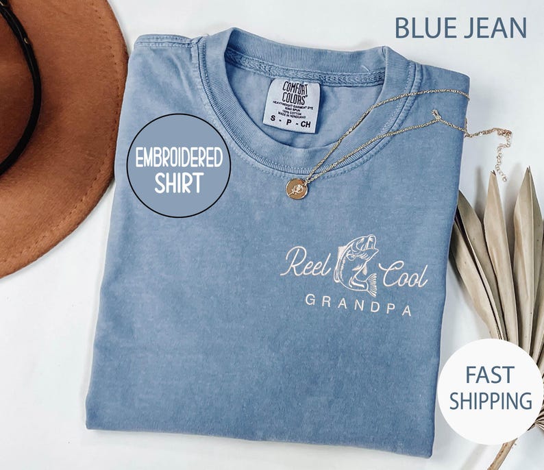 May include: Blue jean colored embroidered shirt with the text "Reel Cool Grandpa" and a fish graphic. The shirt has a round neckline and short sleeves. A gold necklace with a pendant is draped on the shirt.
