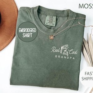 May include: Moss green embroidered shirt with the text "Reel Cool Grandpa" and a fish graphic. The shirt has a crew neck and a small tag on the collar. A gold necklace is draped on the shirt.