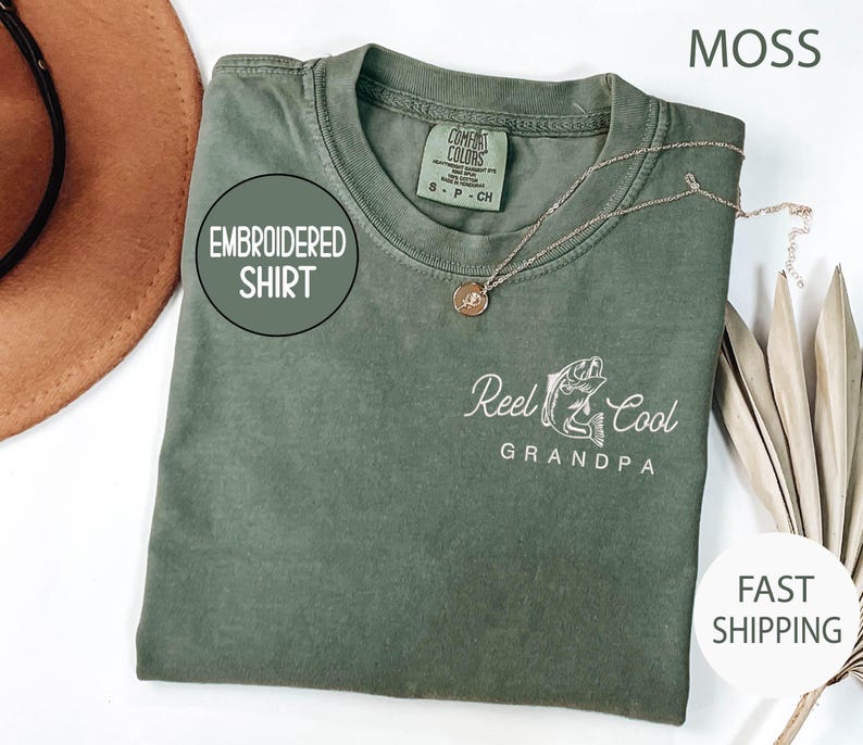 May include: Moss green embroidered shirt with the text "Reel Cool Grandpa" and a fish graphic. The shirt has a crew neck and a small tag on the collar. A gold necklace is draped on the shirt.