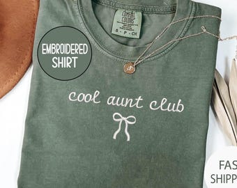Puede incluir: Camiseta verde oliva con el texto bordado "cool aunt club" y un lazo. Un collar dorado con un colgante descansa sobre la camiseta. La camiseta tiene una etiqueta que dice "Comfort Colors".