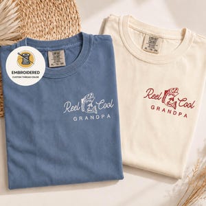 May include: Two t-shirts, one blue and one cream-colored, with the text "Reel Cool Grandpa" embroidered on the front. The blue shirt has white embroidery, while the cream shirt has red embroidery. The shirts are labeled "Comfort Colors" and are size XL-TG-EG.