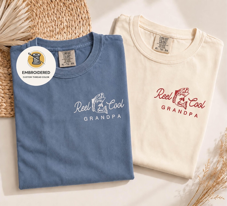 May include: Two t-shirts, one blue and one cream-colored, with the text "Reel Cool Grandpa" embroidered on the front. The blue shirt has white embroidery, while the cream shirt has red embroidery. The shirts are labeled "Comfort Colors" and are size XL-TG-EG.