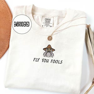 May include: Cream-colored t-shirt with an embroidered wizard design and the phrase "FLY YOU FOOLS" in black. The shirt has a crew neck and short sleeves. A gold necklace with a pendant is draped across the shirt.