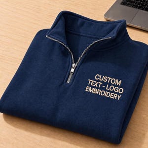 May include: A navy blue quarter-zip pullover featuring a silver-toned metal zipper and the embroidered text CUSTOM TEXT - LOGO EMBROIDERY in a light beige thread on the upper right chest area of the garment.