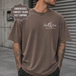 May include: A brown t-shirt with the text "Reel Cool Grandpa" in white, along with a graphic of a fisherman. The shirt also has the text "Embroidered Comfort Colors" in a starburst shape.