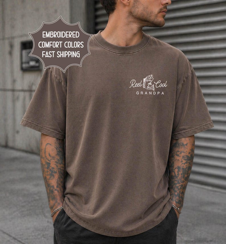 May include: A brown t-shirt with the text "Reel Cool Grandpa" in white, along with a graphic of a fisherman. The shirt also has the text "Embroidered Comfort Colors" in a starburst shape.