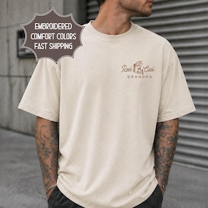May include: Cream-colored t-shirt with short sleeves. The shirt features the text "Reel Cool Grandpa" in brown, along with a graphic of a fish and fishing rod. The shirt also has the text "Embroidered Comfort Colors" on a separate label.