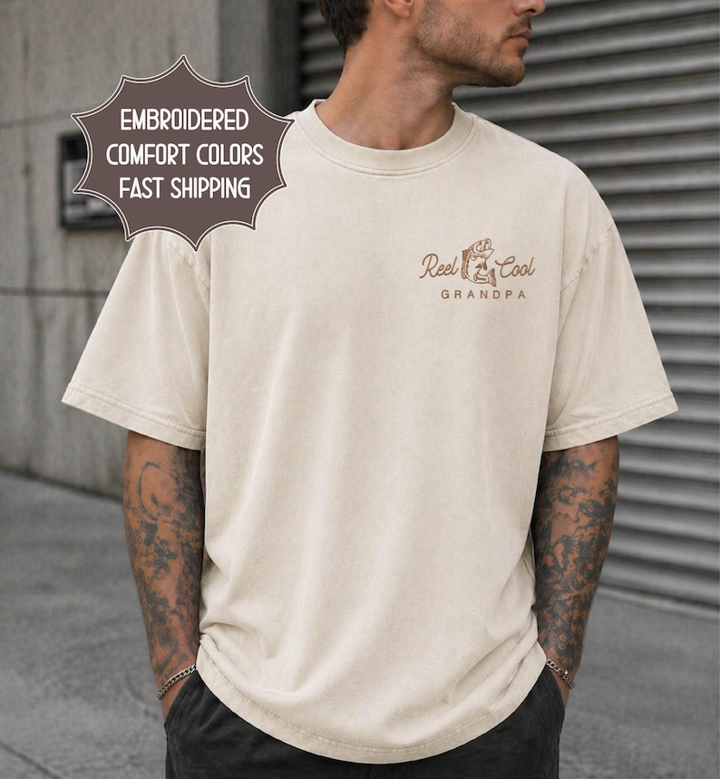May include: Cream-colored t-shirt with short sleeves. The shirt features the text "Reel Cool Grandpa" in brown, along with a graphic of a fish and fishing rod. The shirt also has the text "Embroidered Comfort Colors" on a separate label.