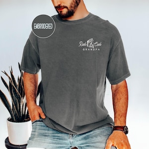 May include: Dark gray t-shirt with the text "Reel Cool Grandpa" and a graphic of a fish and fishing rod. The shirt has a crew neck and short sleeves. The word "Embroidered" is in a circle on the upper left.