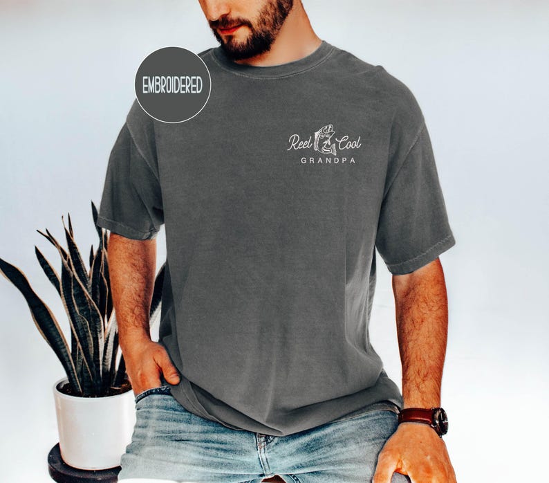 May include: Dark gray t-shirt with the text "Reel Cool Grandpa" and a graphic of a fish and fishing rod. The shirt has a crew neck and short sleeves. The word "Embroidered" is in a circle on the upper left.