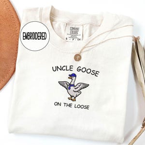 May include: Cream-colored t-shirt with the embroidered text "UNCLE GOOSE ON THE LOOSE" and a cartoon goose wearing a blue hat and scarf. The shirt has a crew neck and short sleeves. A gold necklace with a pendant is draped on the shirt.