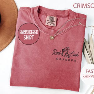 May include: Crimson-colored embroidered shirt with the text "Reel Cool Grandpa" and a fish graphic. The shirt has a crew neck and a small tag on the upper back. A silver necklace with a pendant is draped across the shirt.