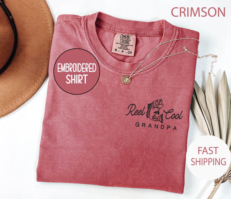 May include: Crimson-colored embroidered shirt with the text "Reel Cool Grandpa" and a fish graphic. The shirt has a crew neck and a small tag on the upper back. A silver necklace with a pendant is draped across the shirt.