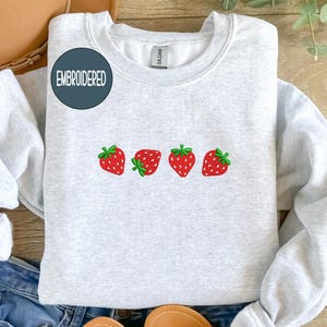 May include: Light gray sweatshirt with four embroidered red strawberries with green stems. The word "EMBROIDERED" is in a navy blue circle on the upper left. The sweatshirt is folded on top of blue jeans.