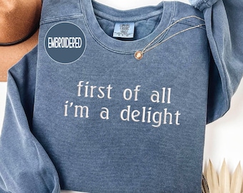 May include: Denim blue embroidered sweatshirt with the text "first of all I'm a delight" in white. A gold necklace with a pendant is draped on the front. The word "DENIM" is at the top.