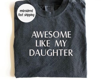 May include: Dark gray t-shirt with the text "AWESOME LIKE MY DAUGHTER" in white. The shirt is folded and has a Comfort Colors tag. A white circle with the text "embroidered fast shipping" is in the upper left corner.
