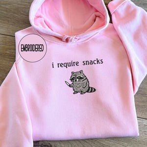 May include: Pink hooded sweatshirt with the text "i require snacks" and an embroidered raccoon graphic holding a knife. The word "EMBROIDERED" is in a pink circle on the upper left. The sweatshirt has a drawstring hood and long sleeves.