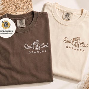 May include: Two folded t-shirts, one brown and one cream-colored, featuring the embroidered text "Reel Cool Grandpa" with a fish graphic. The brown shirt has white embroidery, while the cream shirt has brown embroidery. The shirts are labeled "Comfort Colors" and "XL-TG-EG".