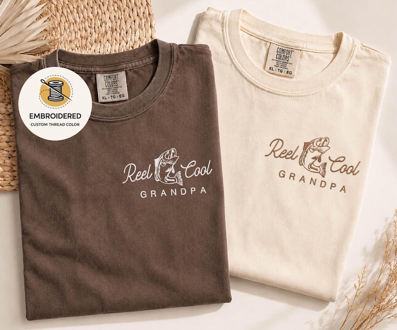 May include: Two folded t-shirts, one brown and one cream-colored, featuring the embroidered text "Reel Cool Grandpa" with a fish graphic. The brown shirt has white embroidery, while the cream shirt has brown embroidery. The shirts are labeled "Comfort Colors" and "XL-TG-EG".