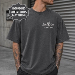 May include: Dark gray t-shirt with the text "Reel Cool Grandpa" and a graphic of a fish and fishing rod. The shirt also has a graphic that says "Embroidered Comfort Colors Fast Shipping". The shirt is a short-sleeved, crew neck style.
