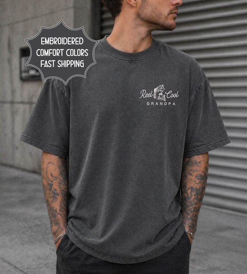 May include: Dark gray t-shirt with the text "Reel Cool Grandpa" and a graphic of a fish and fishing rod. The shirt also has a graphic that says "Embroidered Comfort Colors Fast Shipping". The shirt is a short-sleeved, crew neck style.