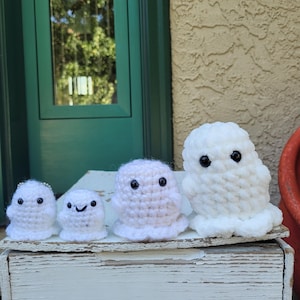 May include: Four handmade crochet ghosts in varying sizes. The ghosts are white with black button eyes and small smiles. The ghosts are displayed on a white, weathered wooden surface, with a green door and stucco wall in the background.
