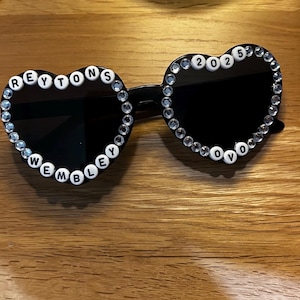 May include: Heart-shaped sunglasses with black lenses and a clear rhinestone border. The left lens spells out "REYTONS WEMBLEY" in white letter beads, and the right lens reads "2025 OVO". The sunglasses rest on a wooden surface.