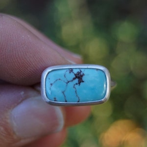 May include: A silver ring with a rectangular turquoise stone set in a bezel setting. The turquoise is a light blue color with brown veins.