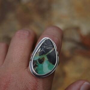 May include: A silver ring featuring a large, teardrop-shaped stone. The stone has a striking pattern of black, green, and brown hues. The ring's setting is a simple, polished silver, highlighting the natural beauty of the stone.