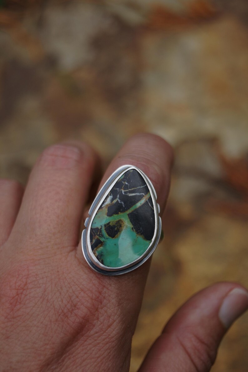 May include: A silver ring featuring a large, teardrop-shaped stone. The stone has a striking pattern of black, green, and brown hues. The ring's setting is a simple, polished silver, highlighting the natural beauty of the stone.