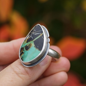 May include: A silver ring featuring a teardrop-shaped stone with a black and green pattern. The stone is set in a silver bezel and has a polished finish. The ring is held up against a blurred background of fall colors.