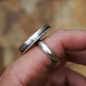 May include: A silver ring with a rectangular top. The ring is made of polished metal and has a simple, modern design. The ring is worn on a finger, showcasing its sleek lines and minimalist aesthetic.