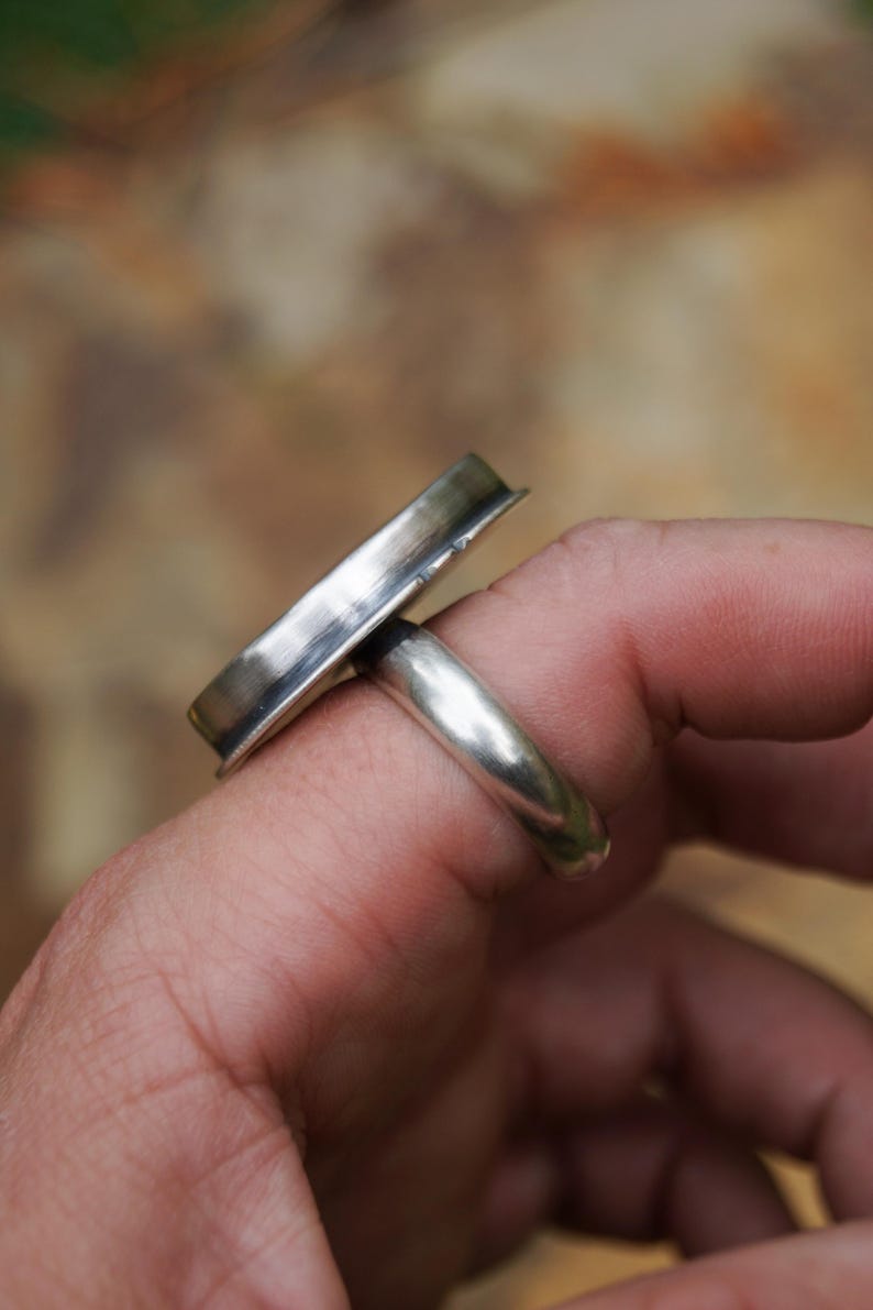 May include: A silver ring with a rectangular top. The ring is made of polished metal and has a simple, modern design. The ring is worn on a finger, showcasing its sleek lines and minimalist aesthetic.
