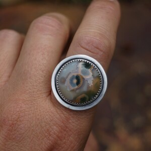 May include: A silver ring with a large, circular stone. The stone displays a unique pattern of green, yellow, and brown hues. The ring is set in a silver bezel with a decorative edge.