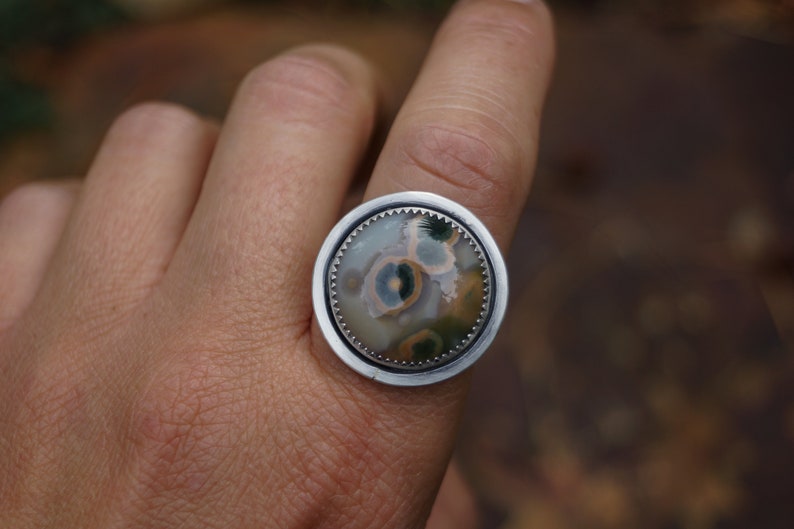 May include: A silver ring with a large, circular stone. The stone displays a unique pattern of green, yellow, and brown hues. The ring is set in a silver bezel with a decorative edge.