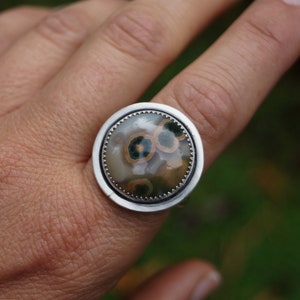 May include: A large, round ring with a silver band. The ring features a polished, circular stone with a pattern of brown, green, and cream-coloured spots. The stone is set in a silver bezel.