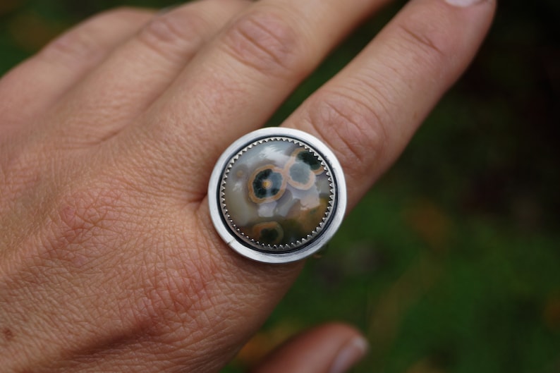 May include: A large, round ring with a silver band. The ring features a polished, circular stone with a pattern of brown, green, and cream-coloured spots. The stone is set in a silver bezel.