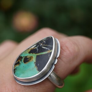 May include: A silver ring featuring a large, teardrop-shaped stone. The stone has a striking pattern of black, green, and brown hues. The ring's setting is a simple, polished silver band, highlighting the unique stone. The ring is a piece of jewelry.
