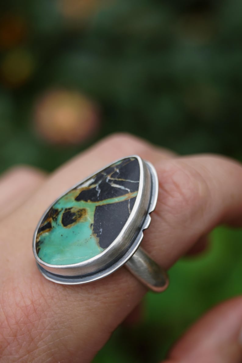 May include: A silver ring featuring a large, teardrop-shaped stone. The stone has a striking pattern of black, green, and brown hues. The ring's setting is a simple, polished silver band, highlighting the unique stone. The ring is a piece of jewelry.