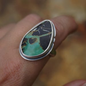 May include: A silver ring featuring a large, teardrop-shaped stone. The stone has a striking pattern of black and green hues, with a polished finish. The ring's setting is a simple, elegant design, showcasing the natural beauty of the stone.