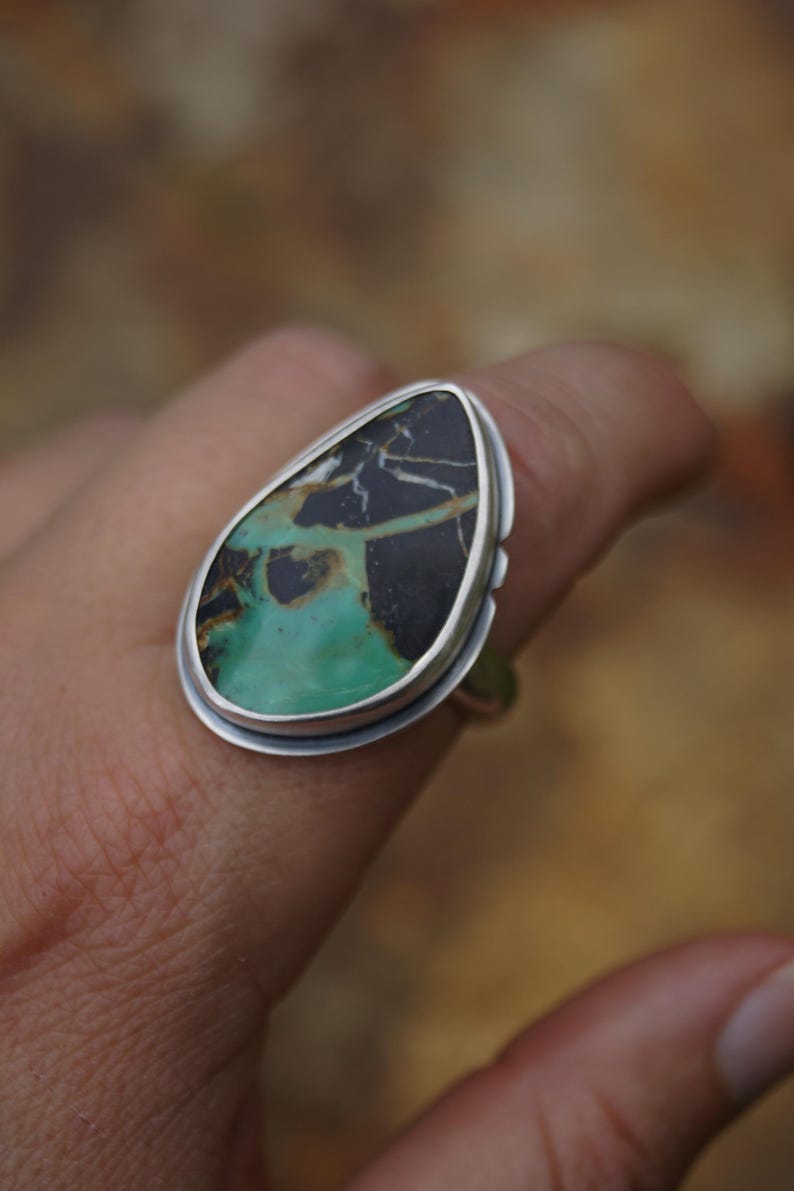 May include: A silver ring featuring a large, teardrop-shaped stone. The stone has a striking pattern of black and green hues, with a polished finish. The ring's setting is a simple, elegant design, showcasing the natural beauty of the stone.