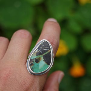May include: A silver ring featuring a large, teardrop-shaped stone. The stone has a striking pattern of black, green, and brown hues. The ring's setting is a simple, polished silver band.
