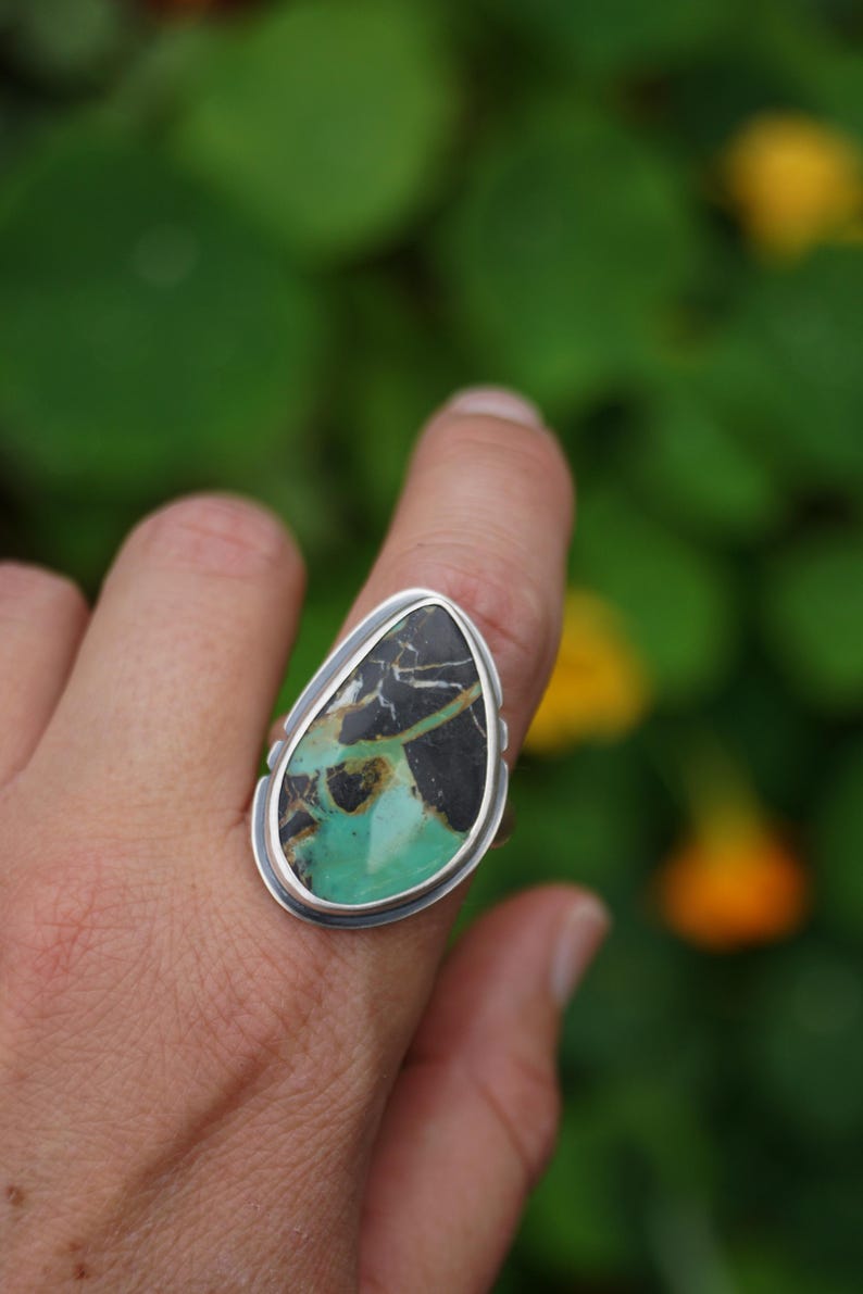 May include: A silver ring featuring a large, teardrop-shaped stone. The stone has a striking pattern of black, green, and brown hues. The ring's setting is a simple, polished silver band.