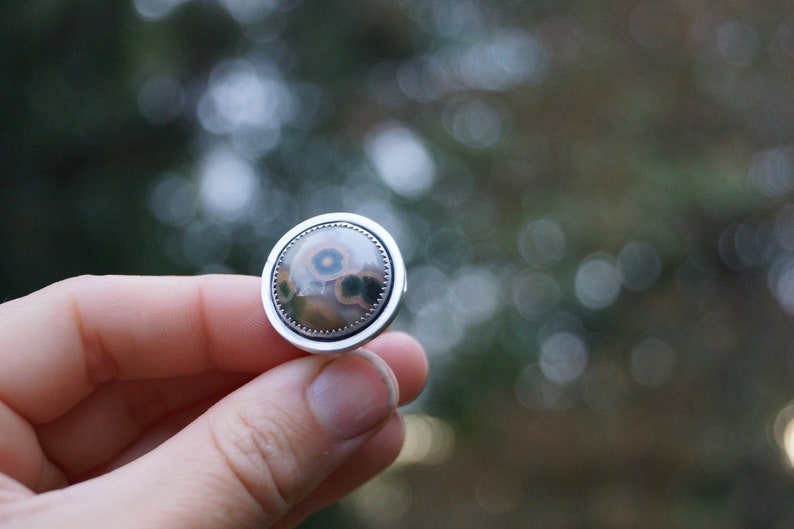 May include: A round, silver cufflink with a polished stone centre. The stone has a unique pattern of brown, green, and blue hues, set within a detailed silver border. The cufflink is held in a hand against a blurred green background.