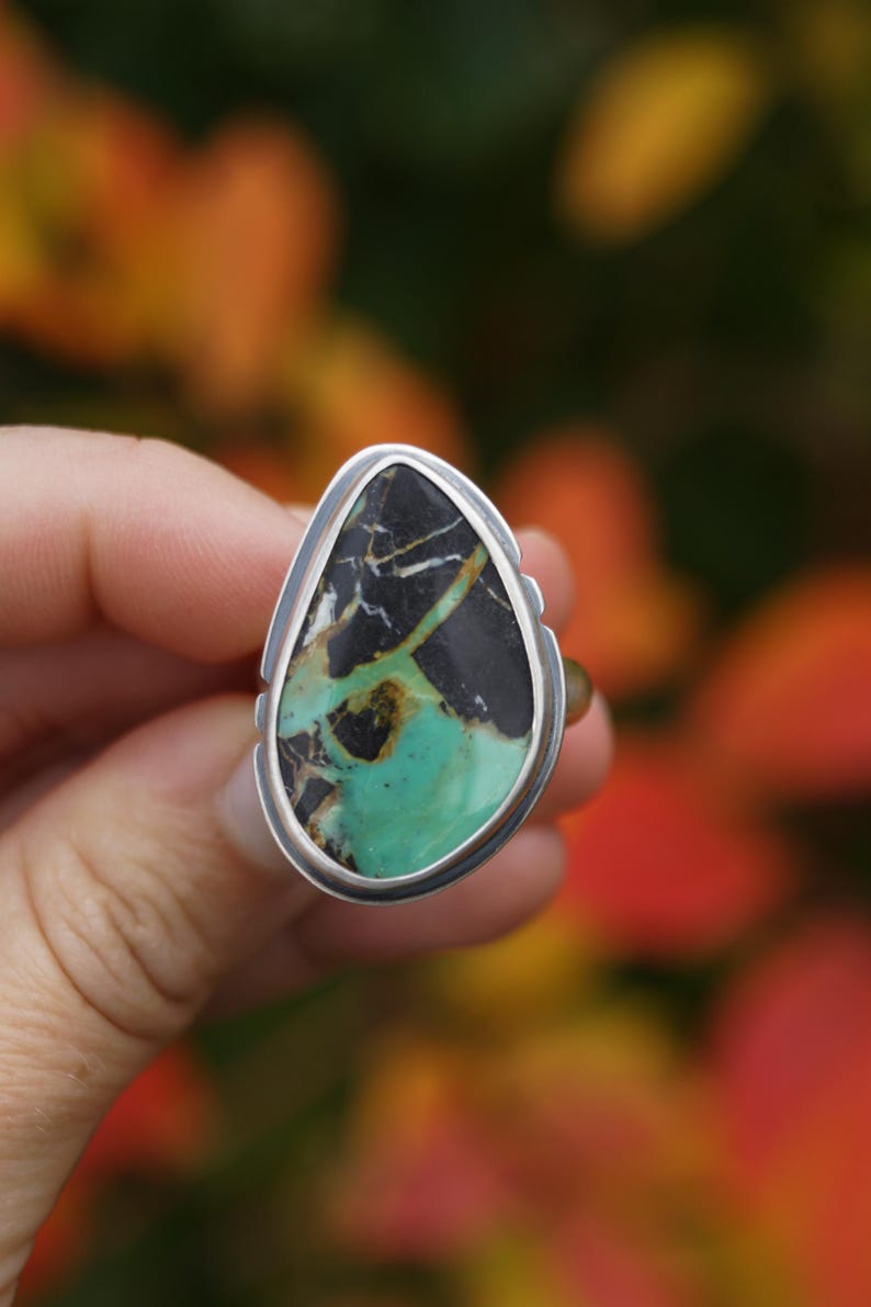 May include: A silver ring featuring a large, teardrop-shaped stone. The stone has a striking pattern of black, white, and turquoise green. The ring is set against a blurred background of fall foliage.